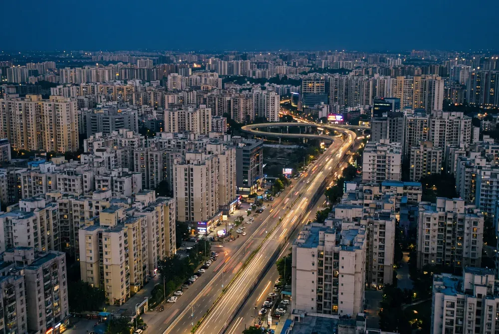 A dense Indian city from the air at blue hour, mid-rise blocks stretching to the horizon with light trails along a broad road.